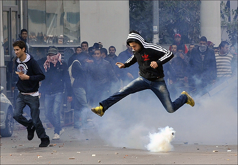 Un manifestant tunisien saute pour éviter les gaz lacrymogènes envoyés par les forces de police pour disperser un rassemblement devant le ministère de l’Intérieur après l’assassinat [9].