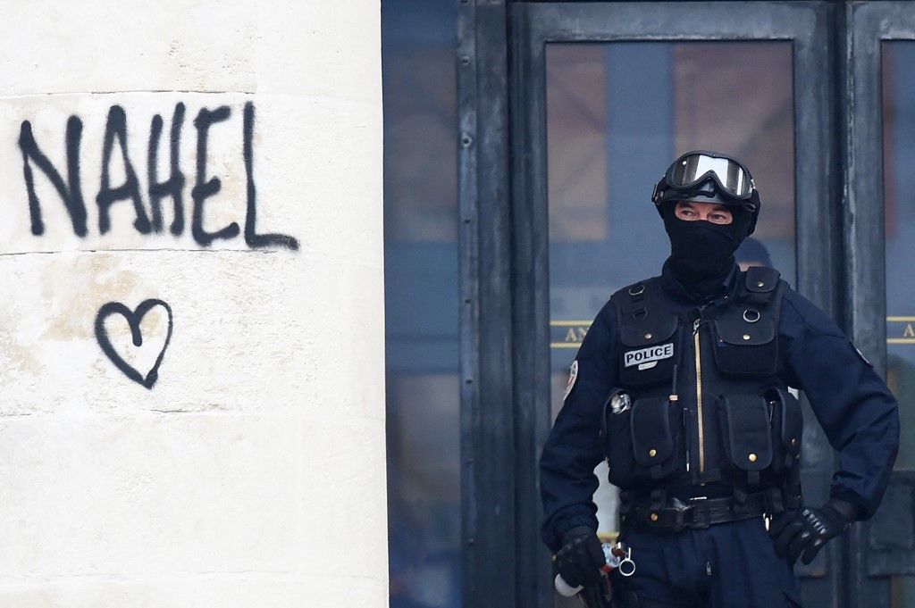 Un mur taggé "Nahel" et, à côté, un policier avec casque et gilet para-balles