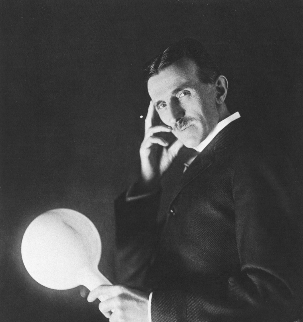 Portrait en noir et blanc de l'inventeur américain Nikola Tesla tenant une lampe allumée dans sa main.