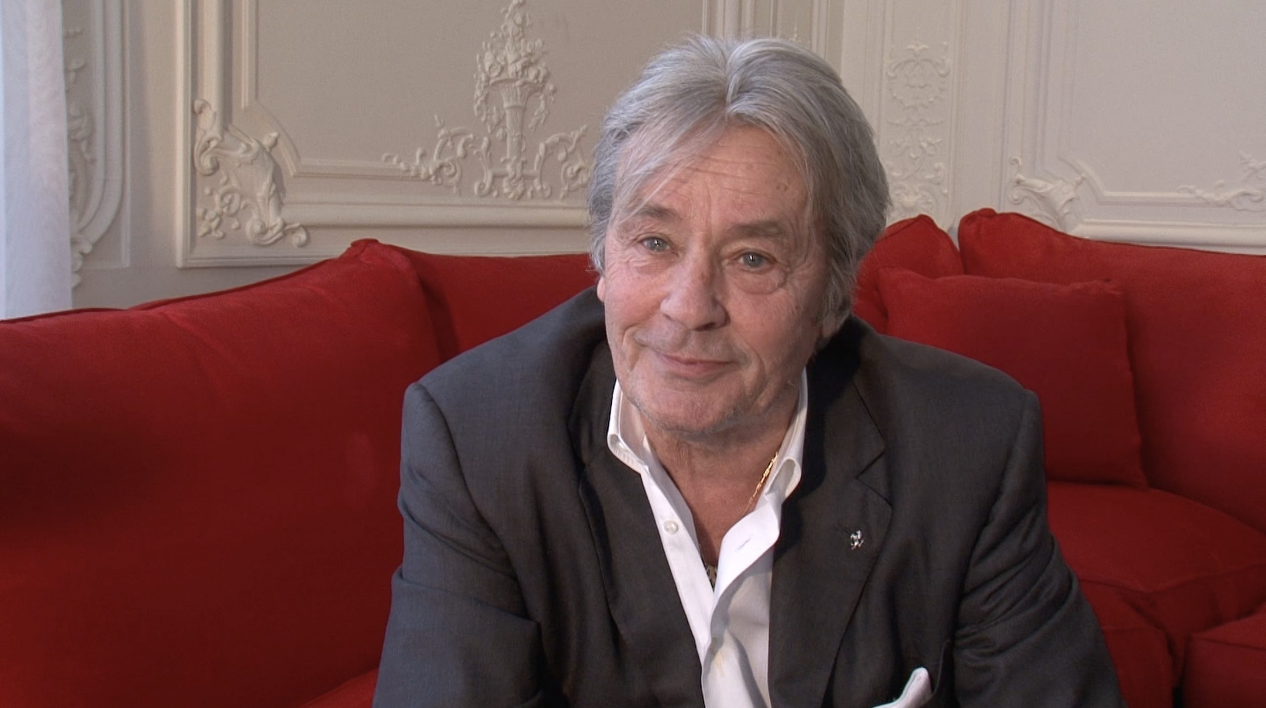 Assis sur un canapé rouge, Alain Delon est interviewé à son domicile.