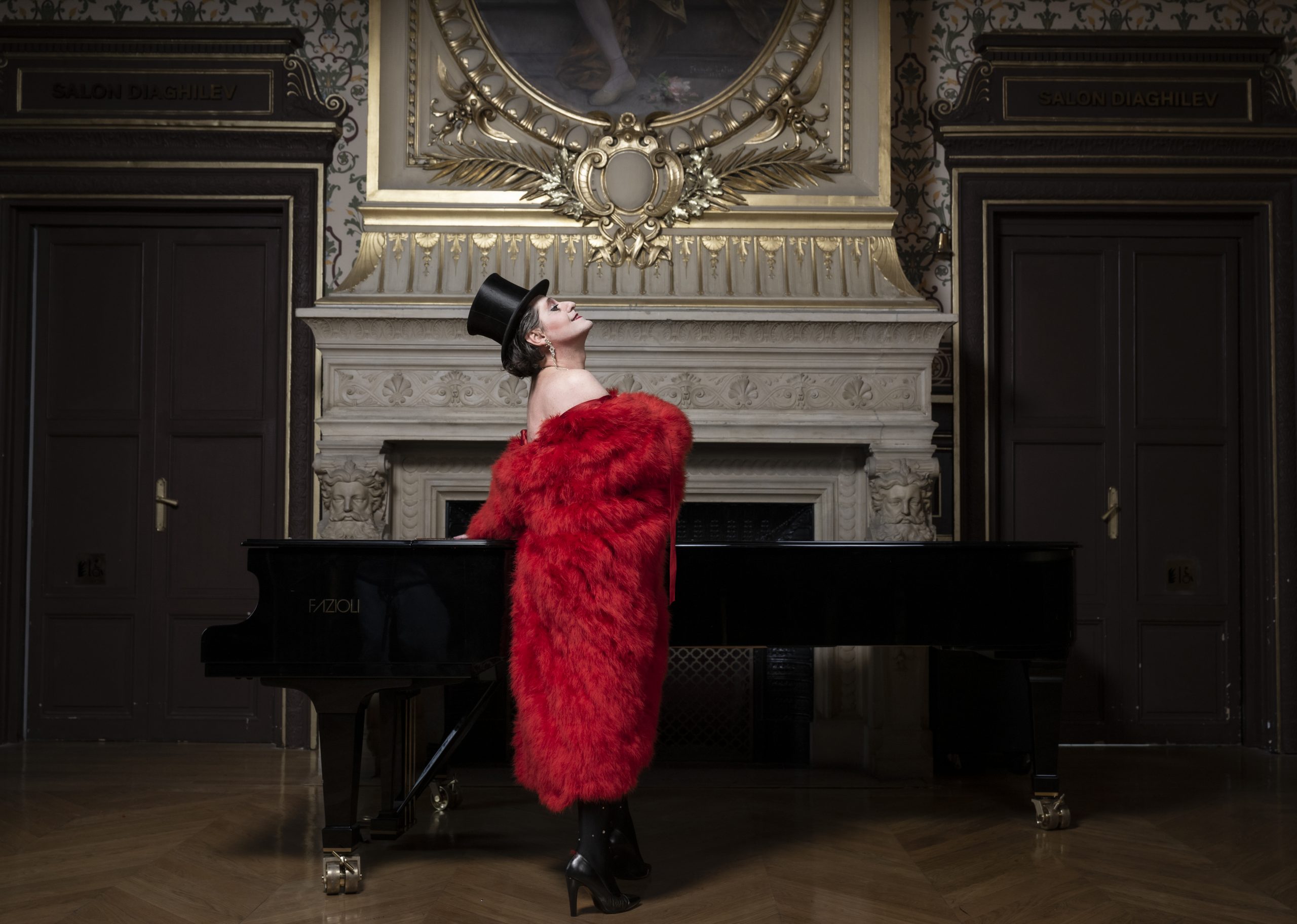 Olivier Py dans le rôle de Miss Knife dans une salle du théâtre du Chatelet. Le comédien porte des talons et un manteau de fourrure rouge, avec un chapeau haut de forme noir. Il est accoudé à un piano à queue.