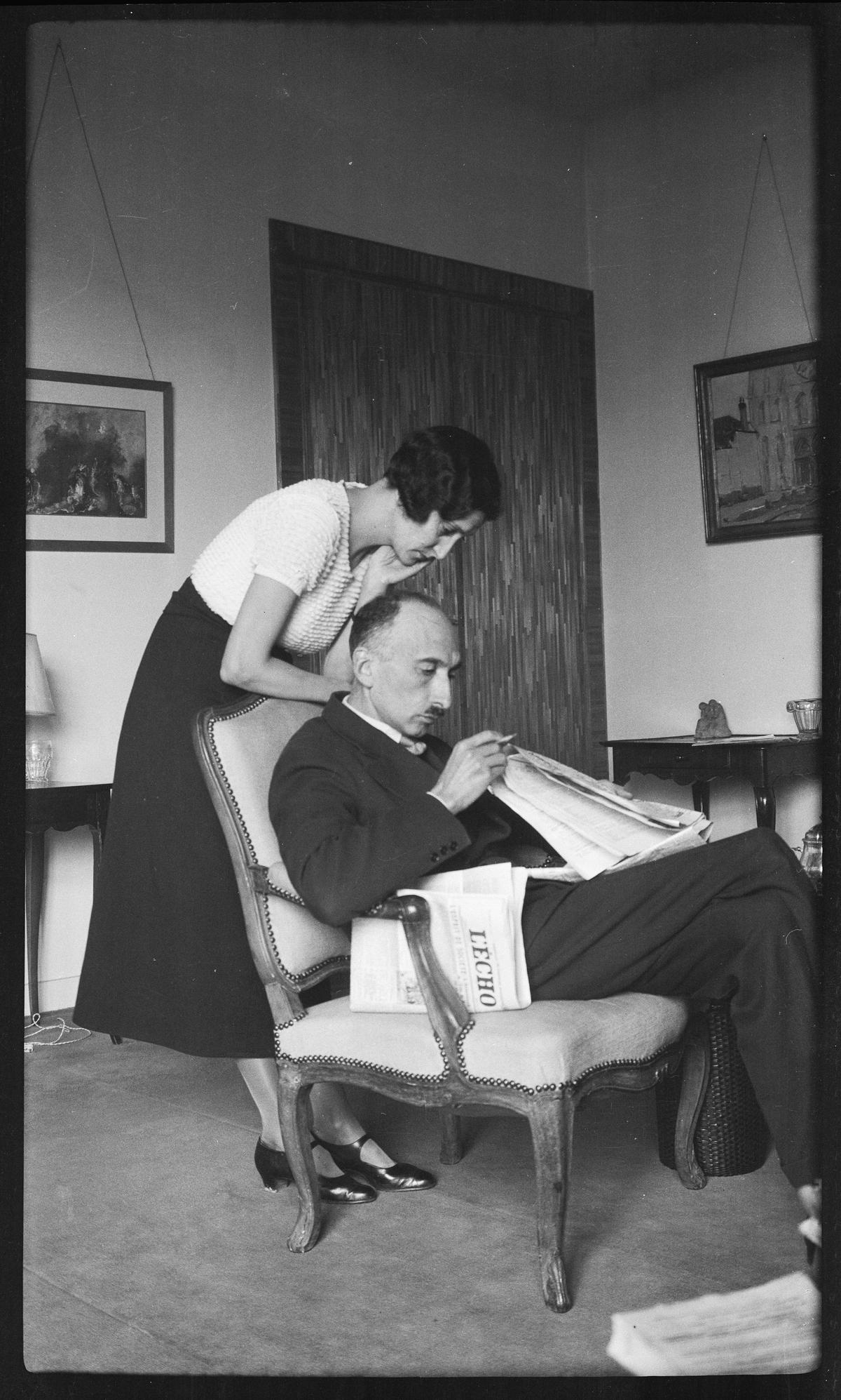 François Mauriac et Jeanne Lafon dans un appartement. François Mauriac est assis avec un journal entre les mains et Jeanne Lafon se tient derrière son fauteuil.