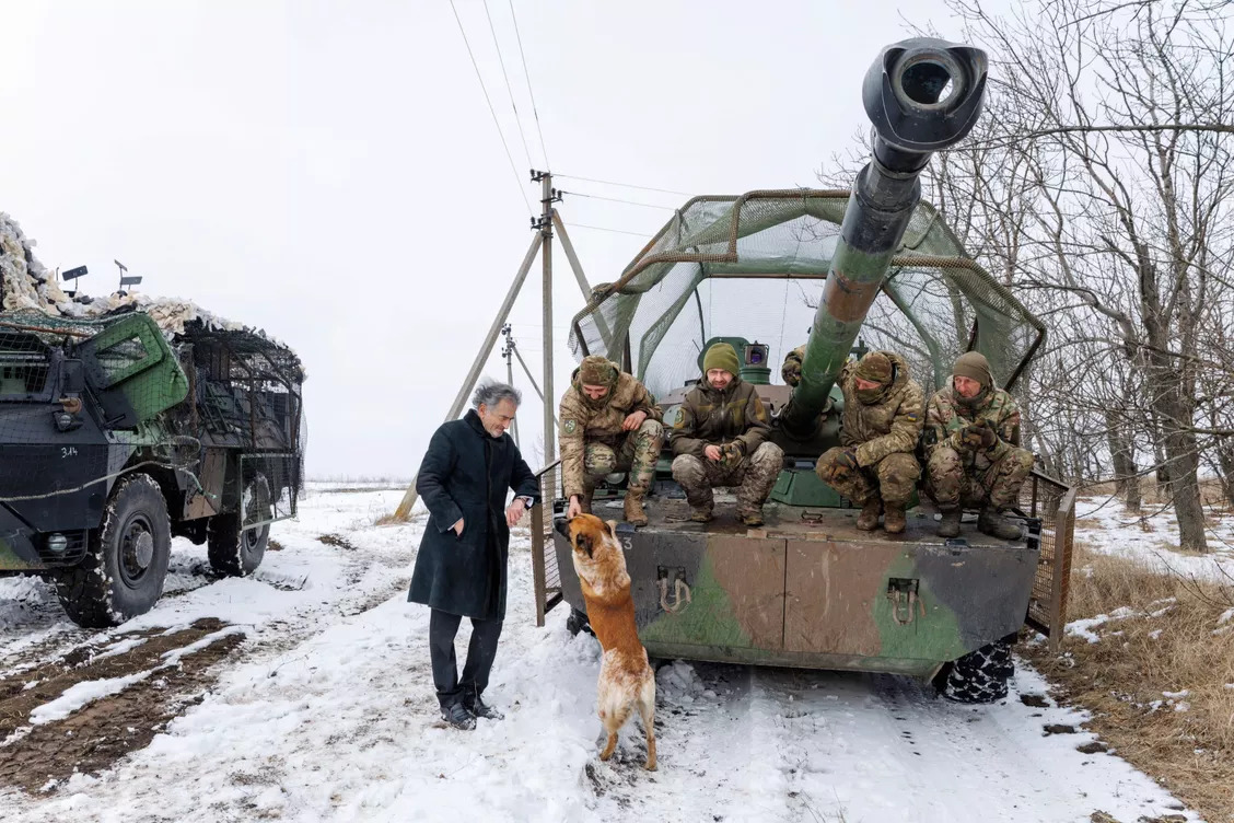 Dans une plaine enneigée dans le Donbass en Ukraine Bernard-Henri Lévy est accoudé à un véhicule blindé. Quatre militaires en tenue de combat sont assis à l'avant du blindé, sous le canon. Au pied du véhicule un chien brun se dresse et regarde le philosophe tout en tendant la patte à un soldat.