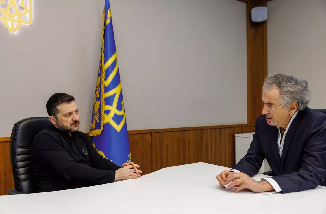 Zelensky et Bernard-Henri Lévy sont assis autour d'une table blanche et parlent dans un bureau austère aux murs blancs. On voit un drapeau jaune et bleu contre le mur.