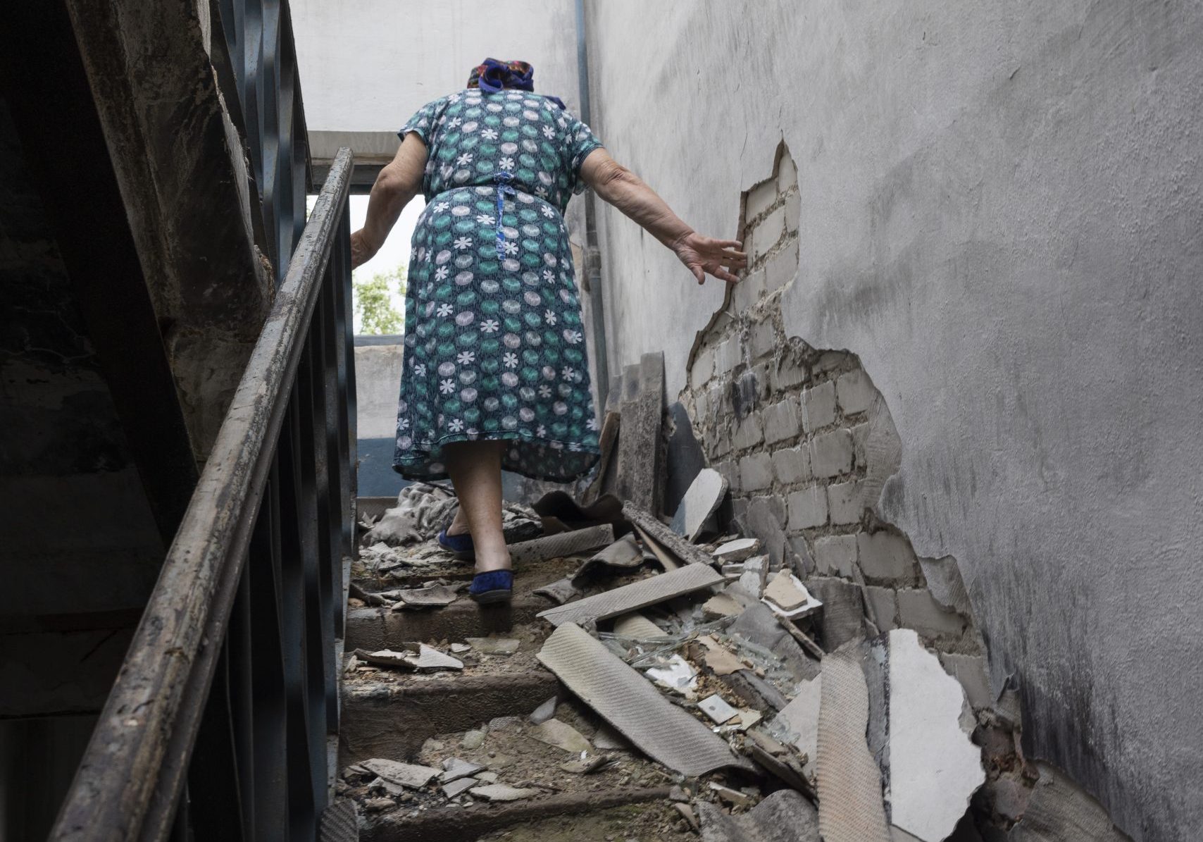 Une vieille dame de dos monte un escalier d'une maison en ruines, détruite par un bombardement russe. L'escalier est couvert de débris, il n'y a plus de fenêtre sur le mur du fond, à droit le mur s'effrite.