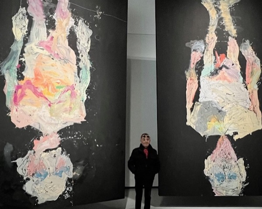 Fernando Arrabal pose entre deux oeuvres monumentales de Georg Baselitz, qui représentent de manière abstraite des corps nus la tête en bas sur des fonds noirs. Arrabal porte des lunettes de vue et a sur le front des lunettes de soleil rondes avec une monture colorée composée de fleurs.