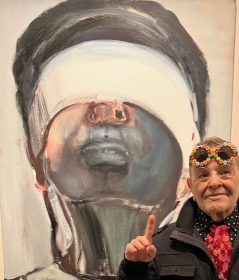 Fernando Arrabal pose à côté d'une toile de Marlene Dumas qui représente un visage avec les yeux bandés, Arrabal pointe le visage du doigt. Arrabal porte des lunettes de vue et a sur le front des lunettes de soleil rondes avec une monture colorée composée de fleurs.