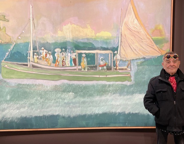 Fernando Arrabal pose devant un oeuvre de Peter Doig, une toile où l'on voit un bateau sur la mer. Arrabal porte des lunettes de vue et a sur le front des lunettes de soleil rondes avec une monture colorée composée de fleurs.