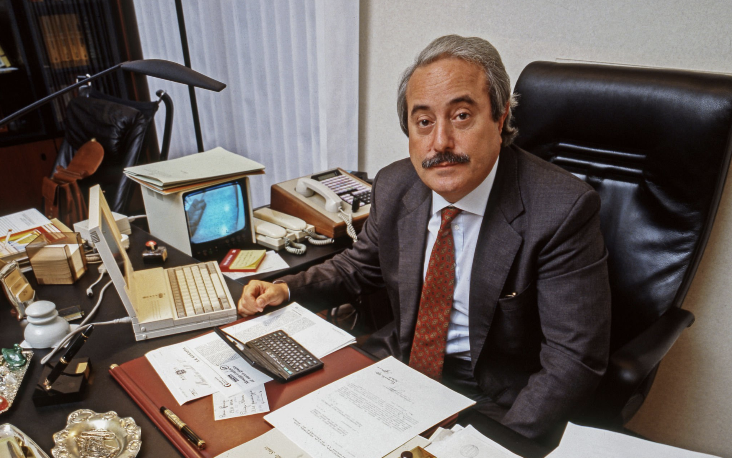 Giovanni Falcone dans son bureau au ministère de la Justice. Le bureau est couvert de papier, il y a deux téléphones et des écrans, un ancien ordinateur des années 90 et une petite télé.