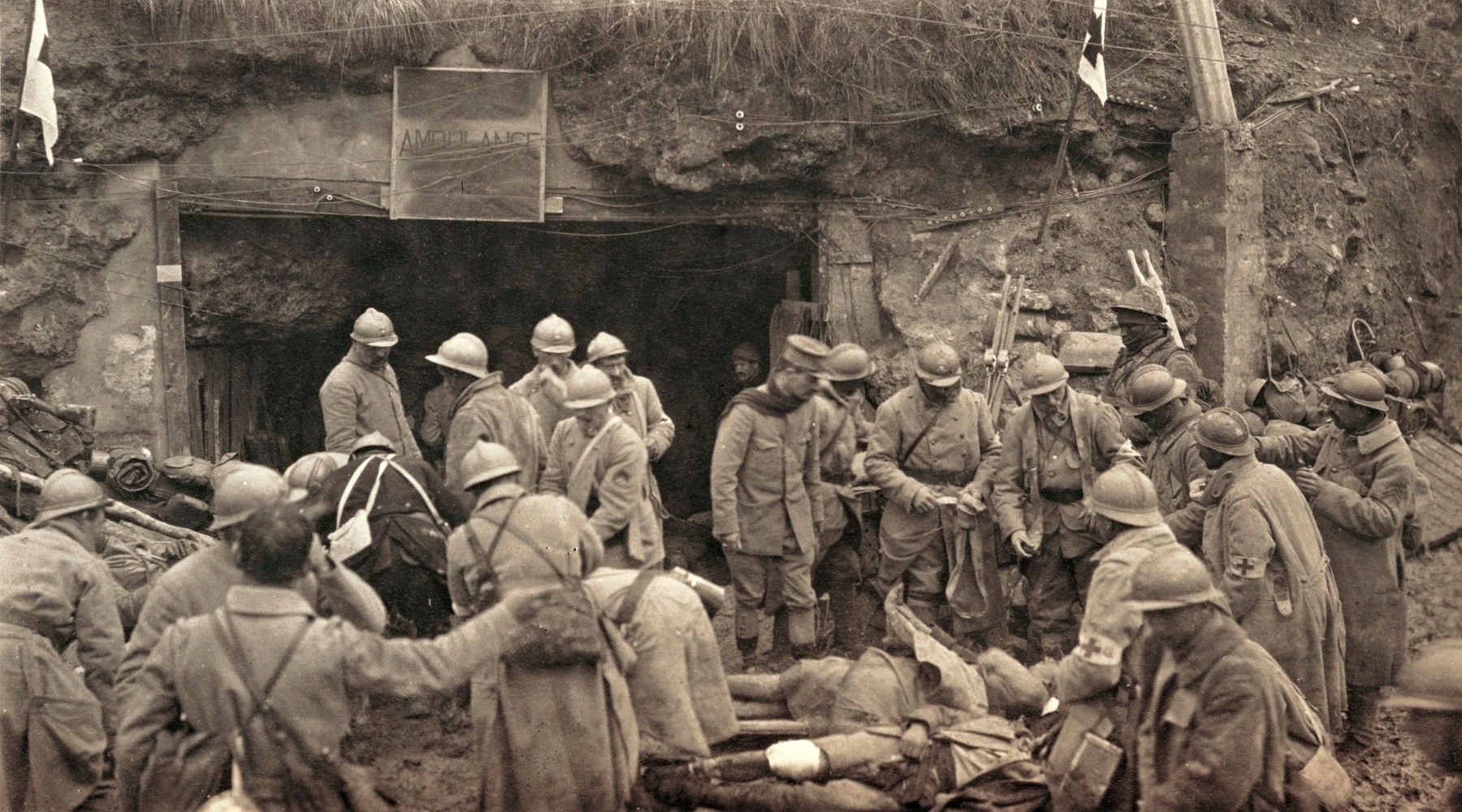 Une trentaine des soldats français de la Première Guerre mondiales, des Poilus, en uniforme, sont rassemblés à un poste de secours. On voit des blessés au sol sur la terre. Le groupe de soldats se tient à l'entrée d'un tunnel souterrains. Au dessus de l'entrée de ce tunnel on voit un panneau avec écrit "Ambulance" et de chaque côté des drapeaux de la Croix-Rouge.