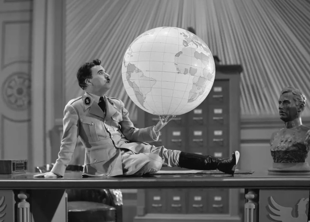 Charlie Chaplin dans le film «Le Dictateur». L'acteur porte un uniforme semblable à ceux d'Hitler, avec une veste croisée et des boittes noires. Il est allongé sur un imposant bureau. Il porte dans sa main un grand globe terrestre et le regarde avec fascination.