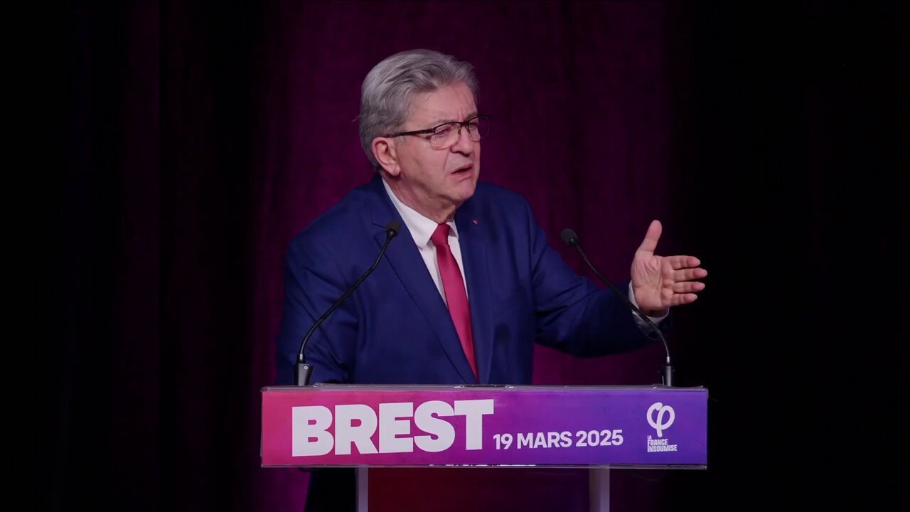 Jean-Luc Mélenchon lors d'un meeting à Brest, le 19 mars 2025. Il porte un costume bleu, une chemise blanche et une cravate rouge. Il parle derrière deux micros dressés sur un pupitre rose et violet sur lequel on peut lire "Brest 19 mars 2025".