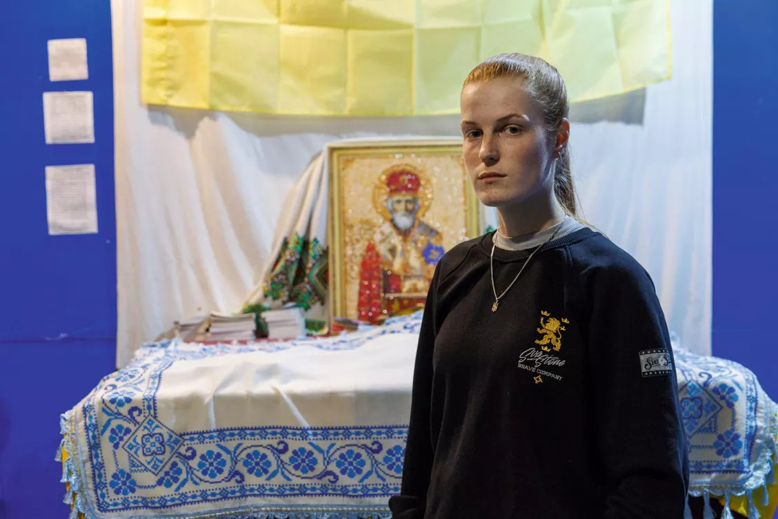 Portrait d'Oksana Rubaniak, jeune femme rousse de 22 ans, commandante de l'unité de drones du 91eme bataillon et poétesse. Elle se tient debout devant un autel religieux orthodoxe un bunker aux murs jaune et bleu.