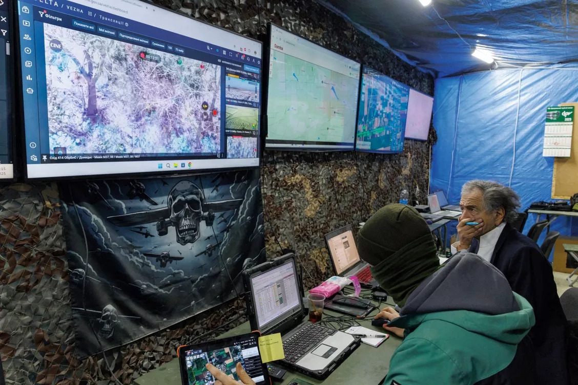 Dans un bunker aux murs recouverts de baches et de filets de camouflage, un soldat cagoulé est assis derrière un bureau lequel sont posés des ordinateurs qu'il regarde. Assis à ses côtés Bernard-Henri Lévy regarde un des 5 écrans accrochés au mur sur lesquels sont retransmis des images filmés par les drones.