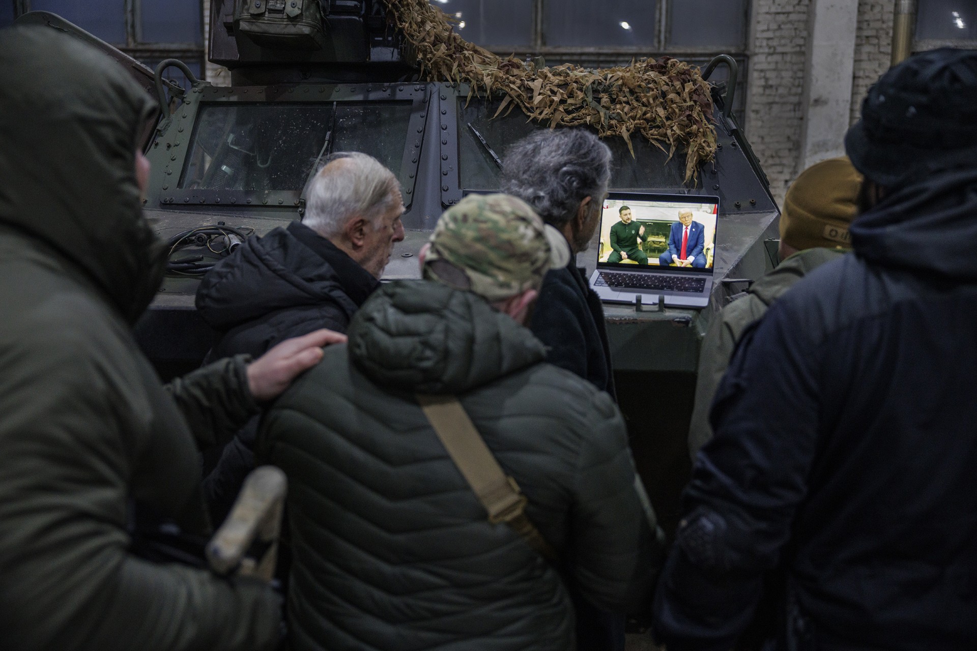 Au premier plan des soldats ukrainiens de dos, en blousons et doudounes kakis. Au second plan, également de dos, on voit Bernard-Henri Lévy et Gilles Hertzog. Tous regardent une vidéo de Zelensky et Trump sur un ordinateur posé sur un char blindé dans un hangar.