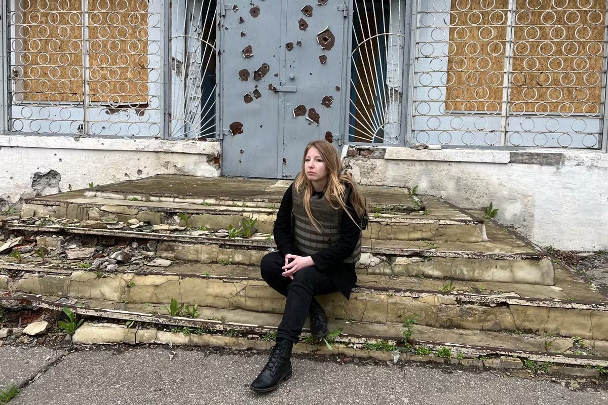 Victoria Amelina avec un gilet parre-balles devant une bibliothèque détruite de la région de Donetsk. Elle est assise sur des marches très abîmées, on voit les impacts de balles sur la porte de l'édifice derrière elle.