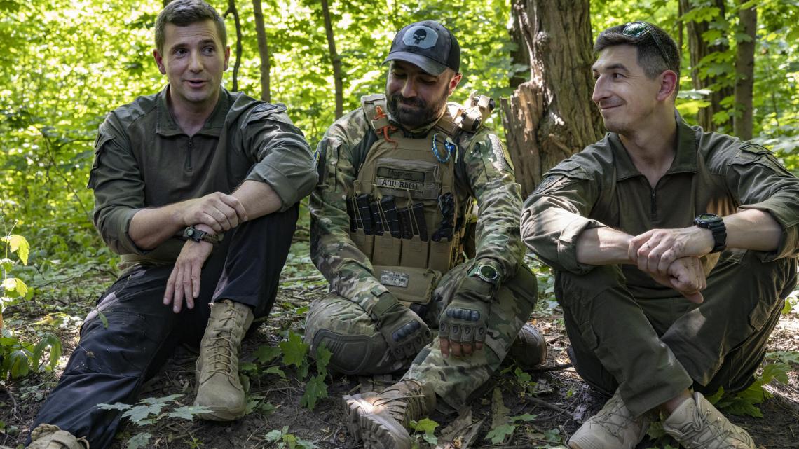 Trois soldats en tenues militaires kaki, treillis, celui au centre porte un gilet-parre-balles avec des chargeurs d'arme automatique. Ils parlent et sont assis au sol dans une forêt.