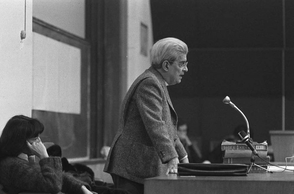Photo en noir et blanc de Jacques Lacan qui donne un séminaire dans un amphithéâtre d'université. Il est debout, de profil, et s'appuie sur un bureau où est posé un micro.