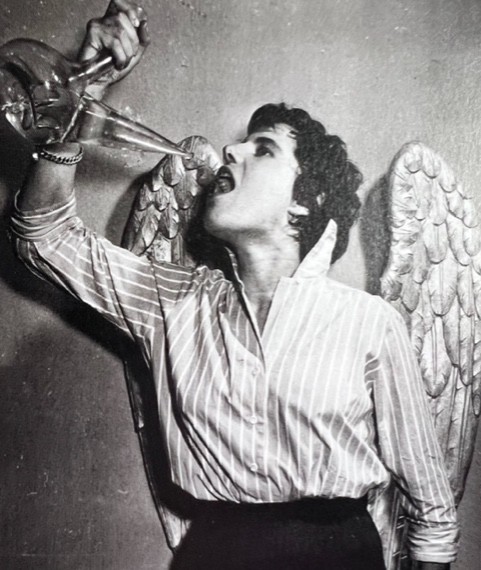 Photo en noir et blanc de Anne Sarraute qui boit à l'aide d'un pichet. Sur le mur derrière elle il y a des ailes d'ange.