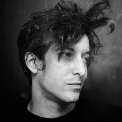 Photo en noir et blanc de Marius Jauffret en costume noir, les cheveux ébouriffés, on distingue de la fumée de cigarette.