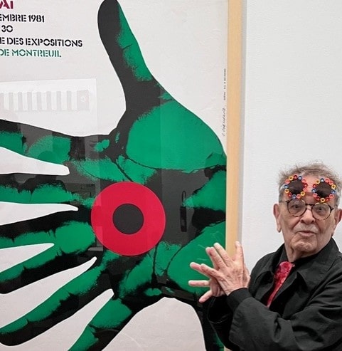 Fernando Arrabal visite l’exposition «Banlieues chéries» au Musée national de l’histoire de l’immigration. Il pose en montrant ses mains devant une affiche où l'on voit une grande main verte avec un rond rouge et noir.