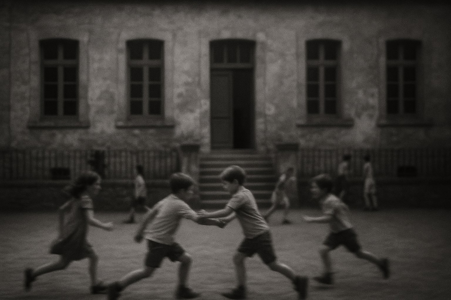 Photographie en noir et blanc, légèrement floue, d’une cour d’école primaire. Des enfants courent et jouent en arrière-plan, tandis qu’au centre de l’image deux garçons se tiennent par le bras.