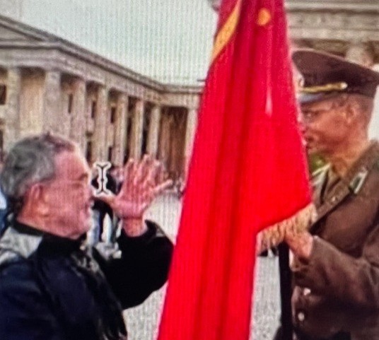 Fernando Arrabal à Berlin, posant sa main ouverte sur le bout de son nez avec un air moqueur, devant un militaire en uniforme portant un drapeau rouge.