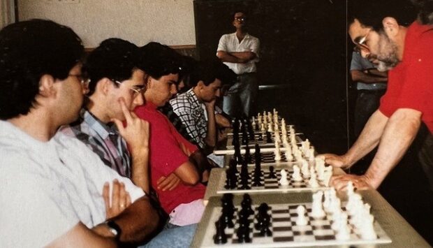 Fernando Arrabal joue aux échecs, debout à droite, face à six joueurs et leurs échiquiers.