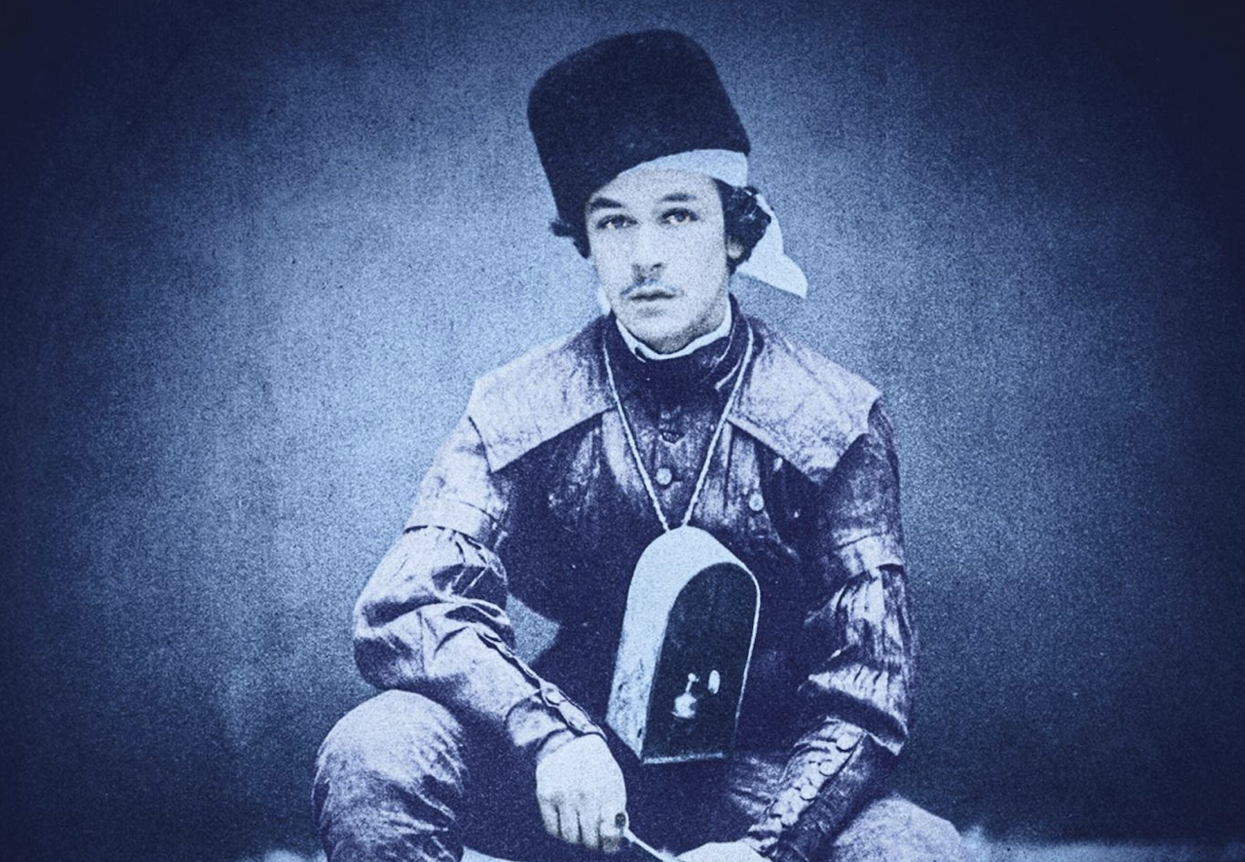 Portrait d’Edmond, l’aïeul de la narratrice dans le roman de Caroline Lamarche : un jeune homme assis, coiffé d’un chapeau, portant un collier orné d’une petite boîte autour du cou. La photographie est dans des tonalités bleutées.