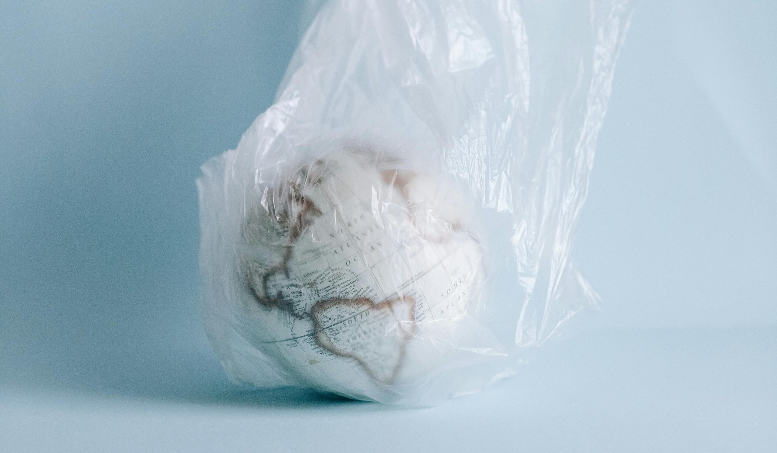 Image en couleur montrant un globe terrestre posé à même le sol, dans un décor blanc et dépouillé. Le globe est entièrement enfermé dans un sac plastique transparent.
