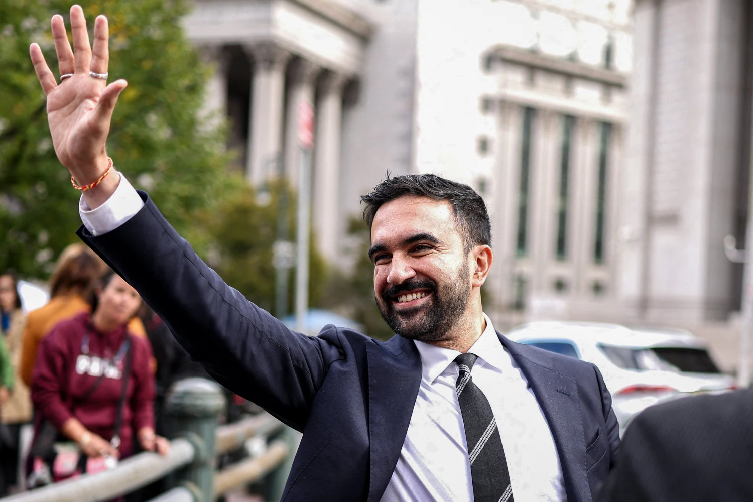 Zohran Mamdani lance un salue en levant la main dans une rue de New York.