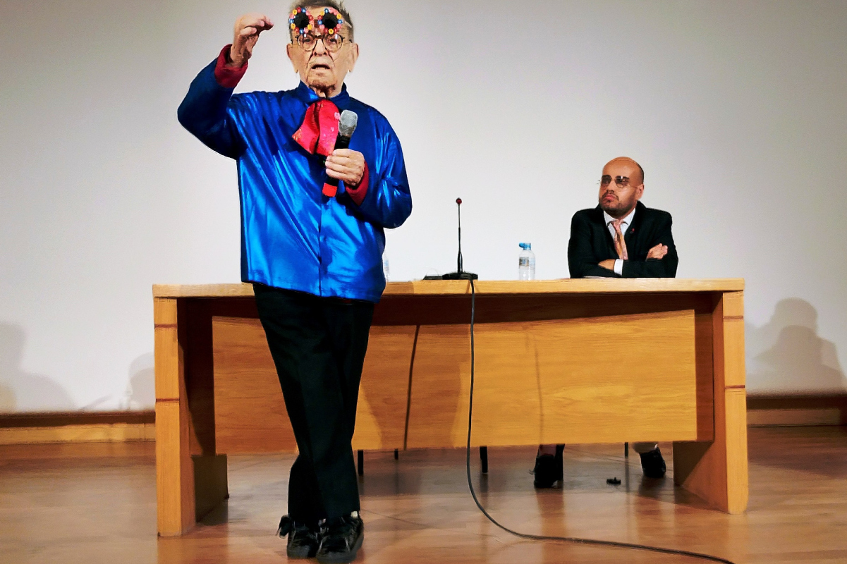 Fernando Arrabal à Don Benito, il parle sur une scène dans un micro, derrière lui il y a un homme assis derrière un bureau.