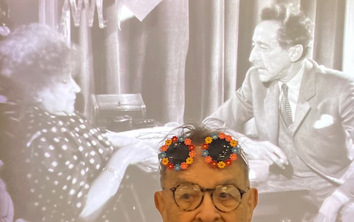 Fernando Arrabal pose devant une image de Colette et Jean Cocteau, on ne voit que le haut du visage d’Arrabal avec ses lunettes colorées.