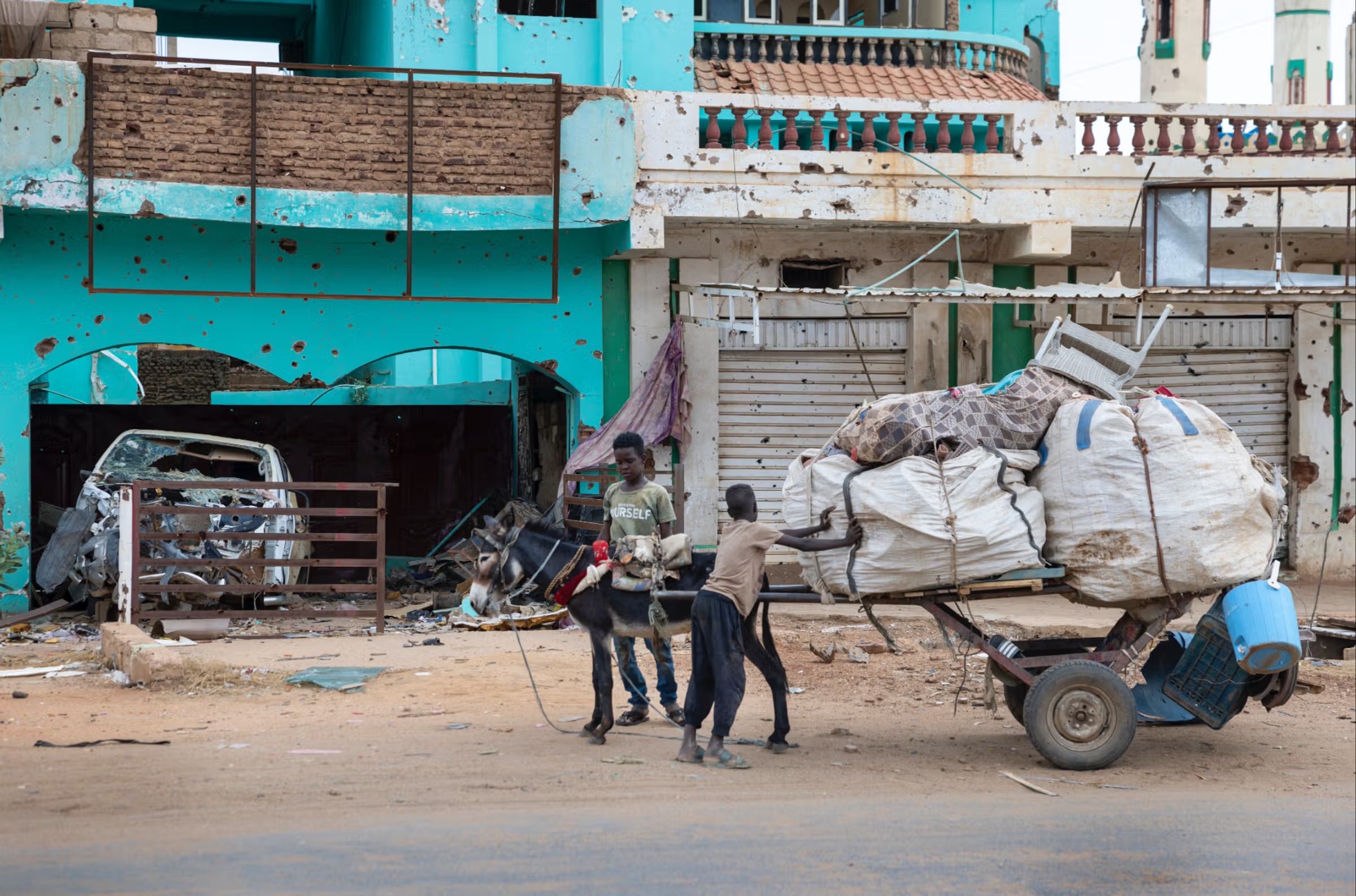 Dans le nord de Khartoum, une famille chassée par les milices rentre chez elle dans une ville où la famine fait des ravages. Deux enfants tirent une charrette couverte de bagages ; derrière eux les murs des maisons sont criblés d'impacts de balles.