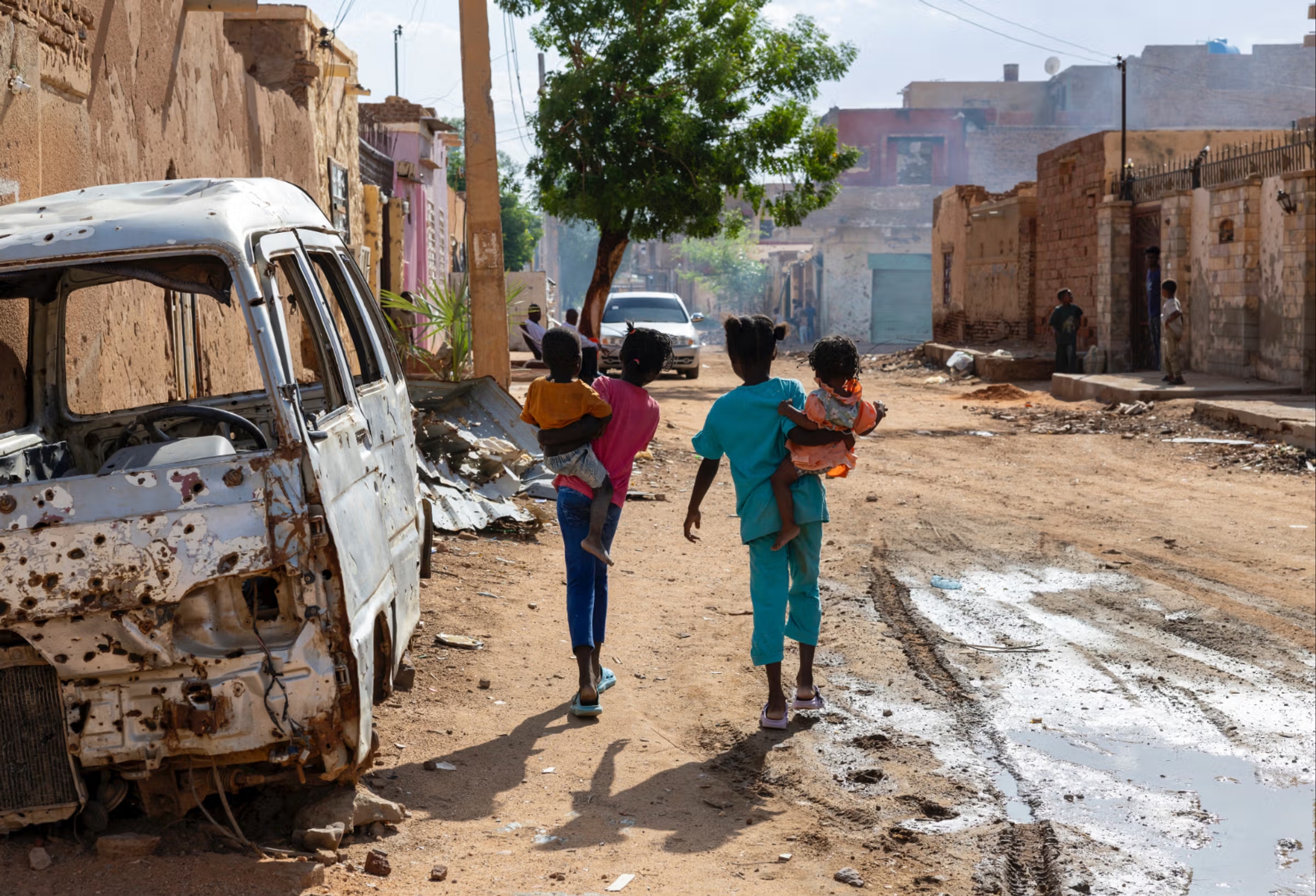 Deux fillettes portent des bébés dans leurs bras dans une rue en terre d'une ville en ruine près d'une carcasse de voiture criblée de balles, dans le quartier de Nubawi, au nord-ouest d’Omdourman.