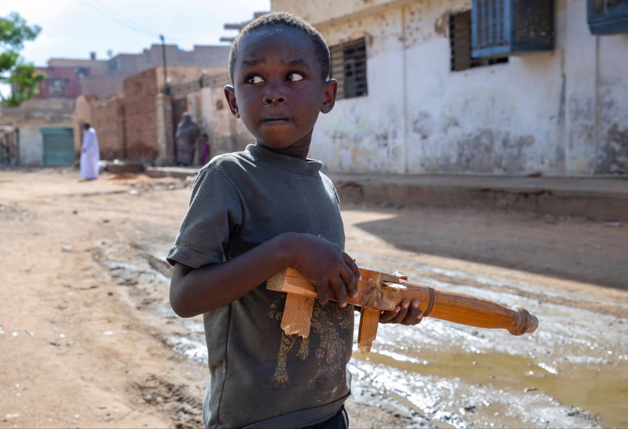 Un enfant au Soudan, dans une rue pauvre, il marche dans la boue et tient un jouer en bois en forme de mitraillette.