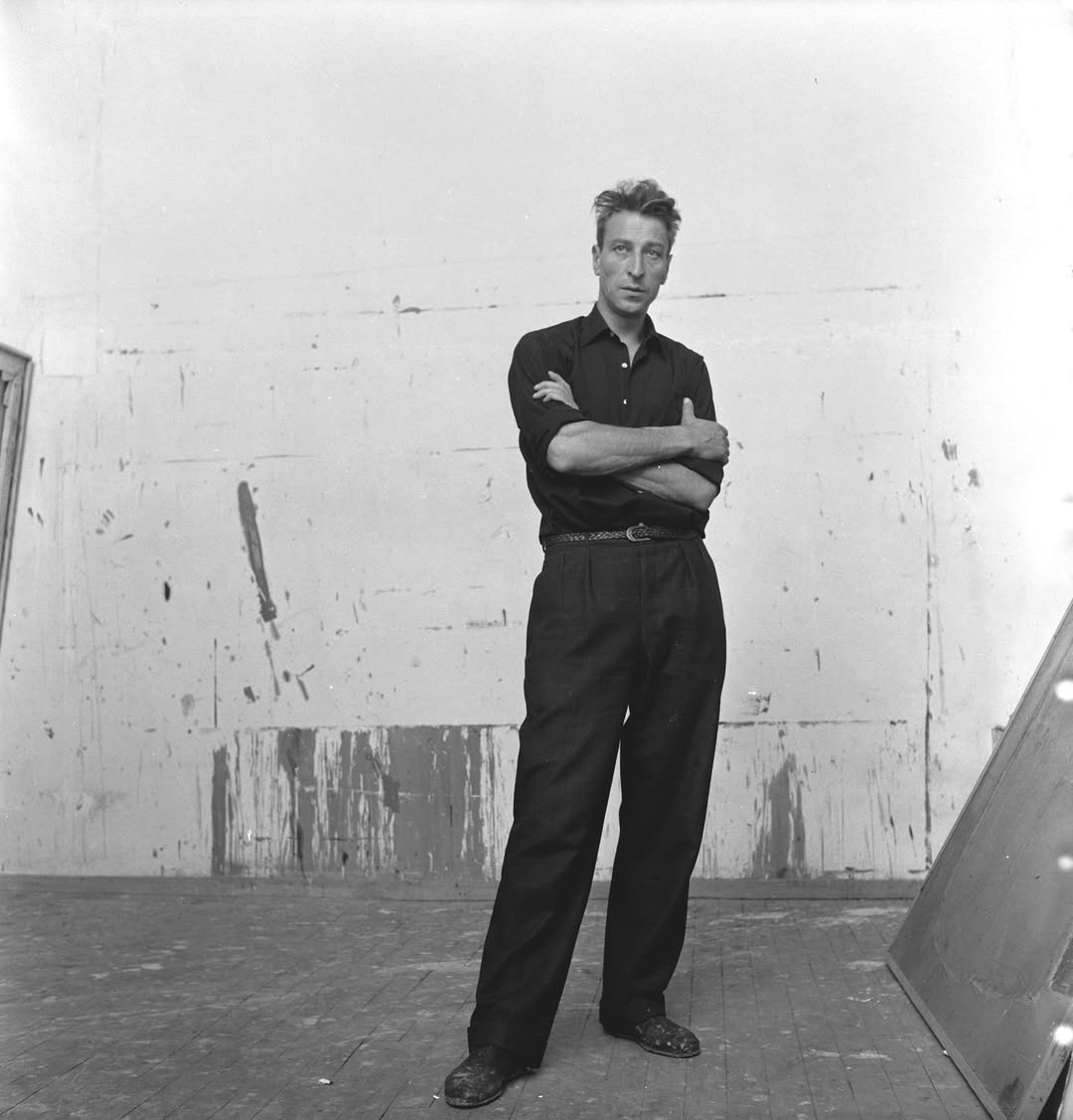 Photo en noir et blanc du peintre Nicolas de Staël habillé en noir, debout, les bras croisés, devant un mur blanc.