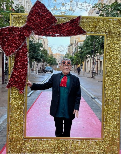 À Melilla, en Espagne, Fernando Arrabal se tient debout dans une rue, sur un tapis rouge, posant derrière un grand cadre doré vide, orné d’un nœud rouge brillant dans l’angle supérieur gauche.