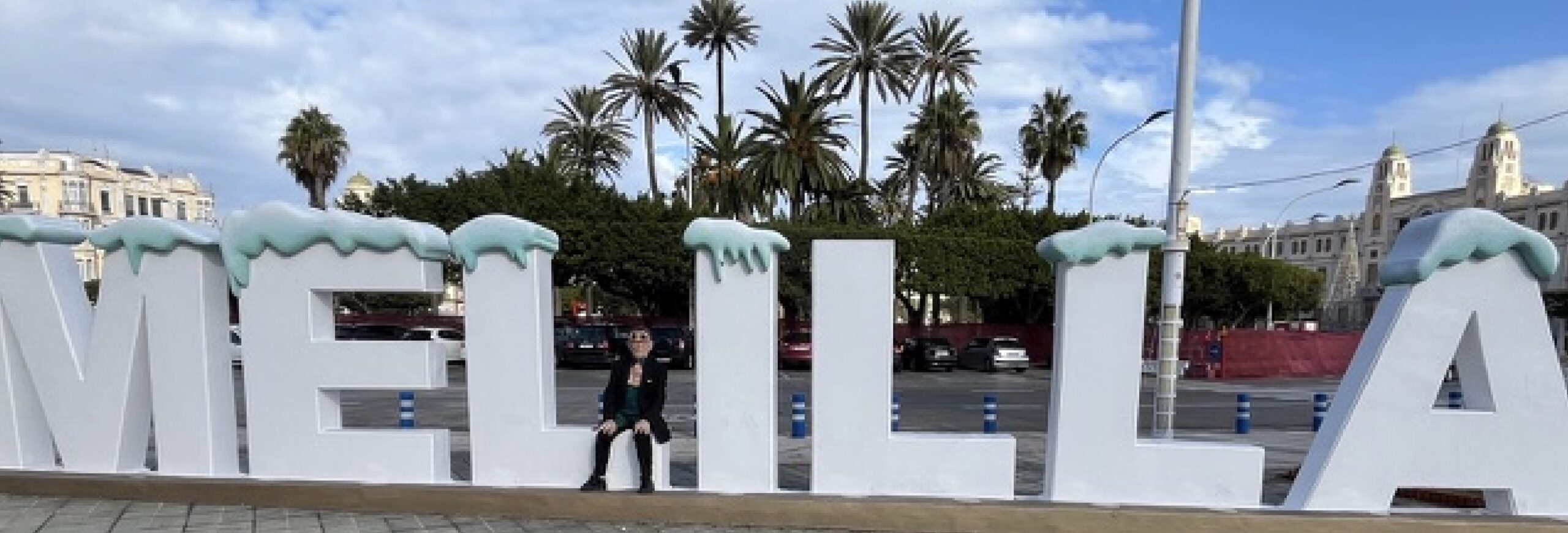 Fernando Arrabal à Melilla, en Espagne, assis sur une sculpture monumentale formant le nom de la ville en grandes lettres blanches.