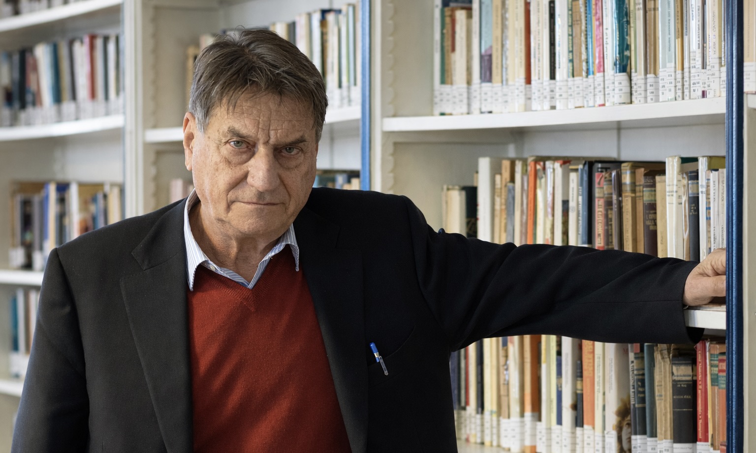 L’écrivain italien Claudio Magris pose dans une bibliothèque, il a le bras posé sur une étagère remplie de livres.