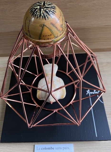 Poème plastique de Fernando Arrabal. Une sculpture montre une colombe blanche enfermée dans une structure géométrique en métal cuivré, surmontée d’un globe ancien. Elle repose sur un socle noir portant le titre « La colombe sans paix. ».