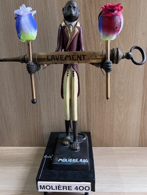 Poème plastique de Fernando Arrabal. Une sculpture représentant un singe anthropomorphe en habit ancien tenant une grande seringue marquée « LAVEMENT », surmontée de deux fleurs colorées, posée sur un socle « MOLIÈRE 400 ».