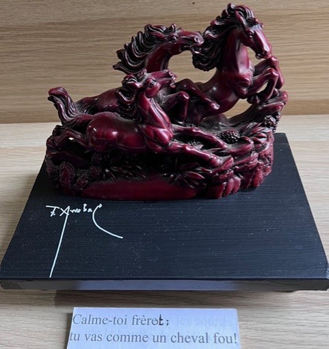 Poème plastique de Fernando Arrabal. Une sculpture en résine bordeaux représentant trois chevaux fougueux en mouvement repose sur un socle noir, avec une inscription (signature d’Arrabal) et un petit texte en dessous.