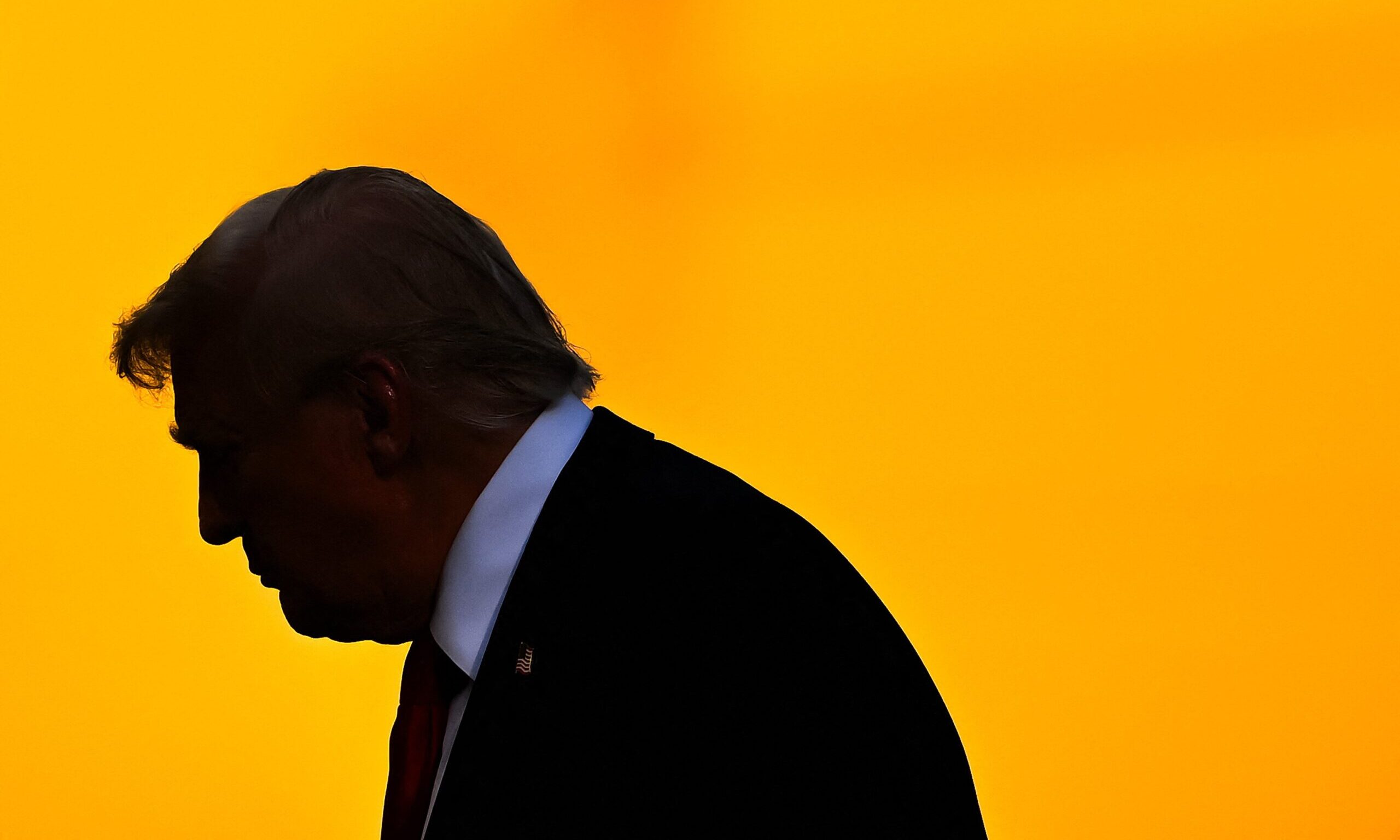 Le président des États-Unis, Donald Trump, de profil, dans l'ombre, devant un fond jaune.