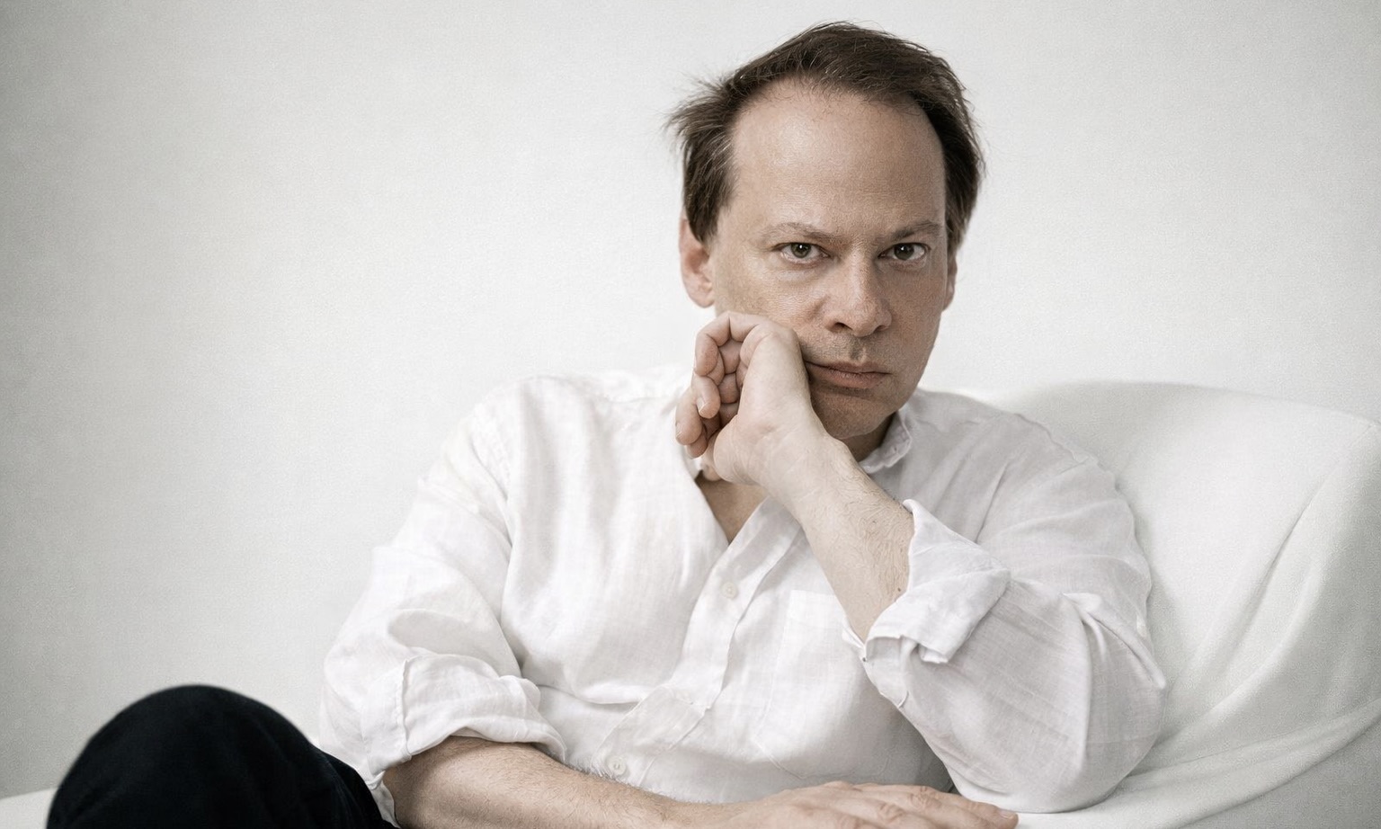 Photo de L’écrivain et essayiste américain Adam Gopnik, assis dans un fauteuil blanc devant un mur blanc.