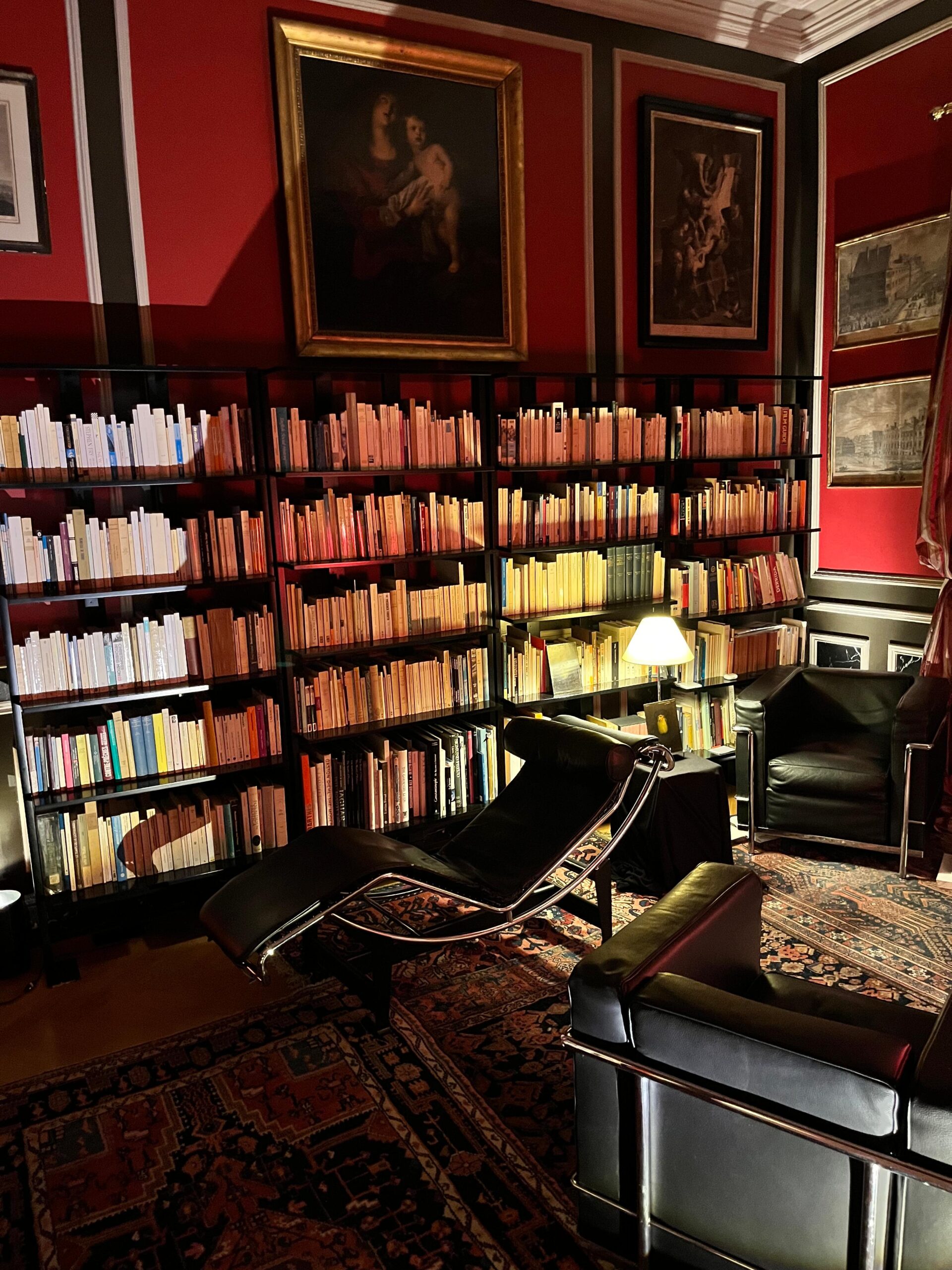 Cabinet de psychanalyse de Michael Larivière. On voit une chaise longue près d'une bibliothèque et deux fauteuils, sur les murs rouges il y a des tableaux.