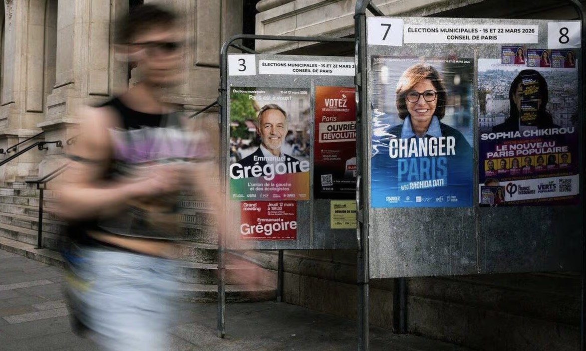 Une personne marche devant des panneaux électoraux installés devant la mairie du 10e arrondissement de Paris, où sont affichées les campagnes de la candidate de LFI Mme. Chikirou, de la candidate de droite Rachida Dati (deuxième à droite) et du socialiste Emmanuel Grégoire (à gauche), quelques jours avant le premier tour des élections municipales du 15 mars 2026, à Paris.