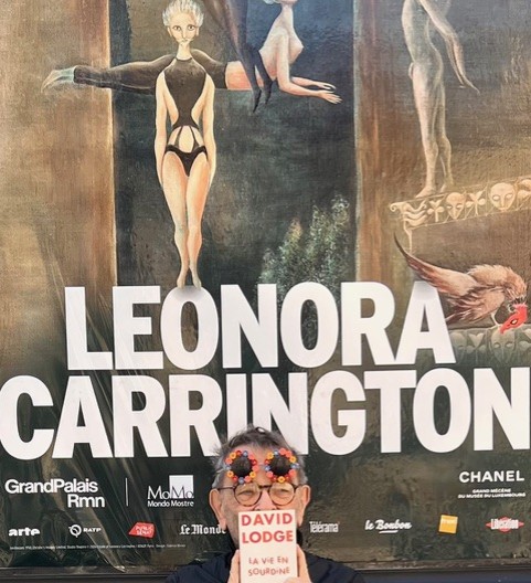 Fernando Arrabal visite l’exposition consacrée à Leonora Carrington au Musée du Luxembourg. Il pose devant l'affiche de l'exposition.