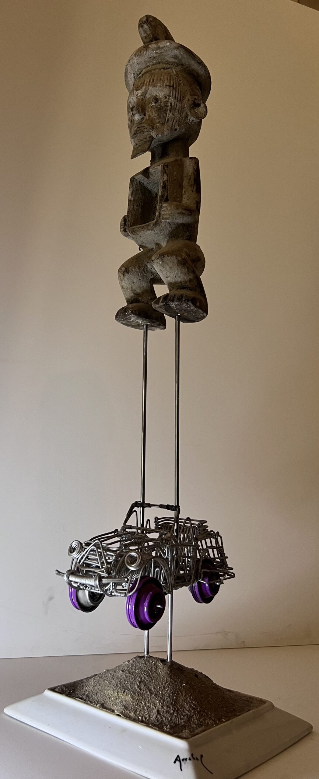 Poème plastique de Fernando Arrabal Une statuette tribale en bois semble flotter au-dessus d’une petite voiture en fils de fer aux roues violettes, posée sur un socle sableux.