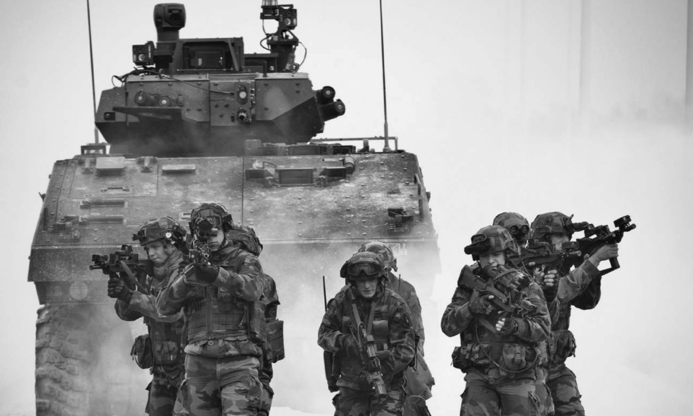 Photo en noir et blanc d'un entraînement de l’armée de terre française. On voit des militaires avec des armes et uniformes marcher devant un char.
